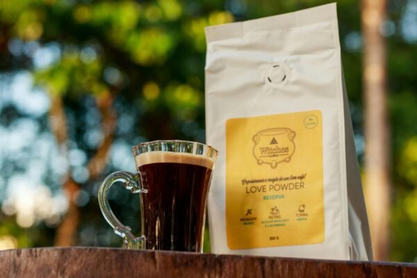 A flavorful coffee drink sits beside a Love Powder coffee bag on a wooden surface amidst a lush green backdrop, evoking freshness.